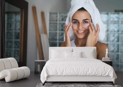 Young smiling female with towel on head puts moisturizing cream on face while standing at home bathroom. 30 years old happy woman doing daily morning rituals. Enjoying healthy skin with collagen care` Wall mural